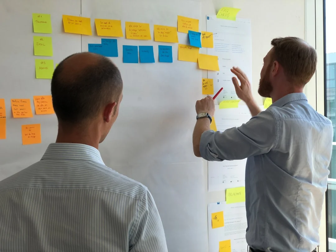 Two businessmen analyze stock market data using a smartphone and laptop in a modern office setting.